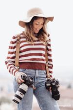Photographer wearing the Brown Vintage leather camera harness over a striped sweater and wide-brimmed hat, holding two Canon cameras with telephoto lenses at hip level, showing the harness straps with D-rings and snap hooks in use.