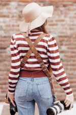Rear view of a photographer wearing the Brown Vintage leather camera harness, showing the X-shaped crossback design with center rivet, adjustable buckles, and brown leather keeper loops over a striped sweater, with two Canon cameras hanging at each side.