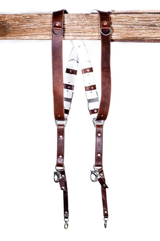 Brown Vintage White Padding leather camera harness displayed on weathered wood beam, showing full X-harness design with brown leather shoulder straps, white padded center strap with brown leather accent bands, metal D-ring connectors, and carabiner clips