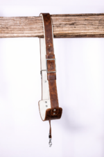 Brown vintage leather camera strap with white padded underside, adjustable buckle, and silver clasp hardware hanging from a rustic wooden beam on a light gray background.