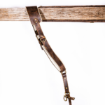 Full-length brown vintage leather camera strap with white shoulder padding, brass-tone rivets and rings, adjustable strap holes, and clasp attachment hanging from a rustic wooden beam on a light gray background.