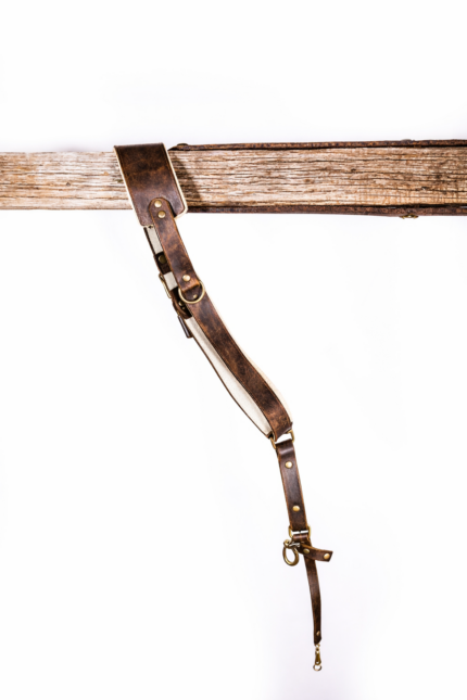 Full-length brown vintage leather camera strap with white shoulder padding, brass-tone rivets and rings, adjustable strap holes, and clasp attachment hanging from a rustic wooden beam on a light gray background.