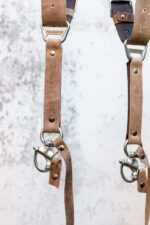 Bottom attachment detail of Brown Wild West Vintage harness showing tan leather strap, metal carabiner clip, brown leather attachment tag with rivet, metal hardware, and engraved connector plate