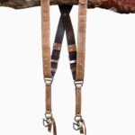 Brown Wild West Vintage leather camera harness displayed on weathered wood log, showing full X-harness design with tan leather shoulder straps, black textured center strap with brown leather accent bands, metal D-ring connectors, and carabiner clips