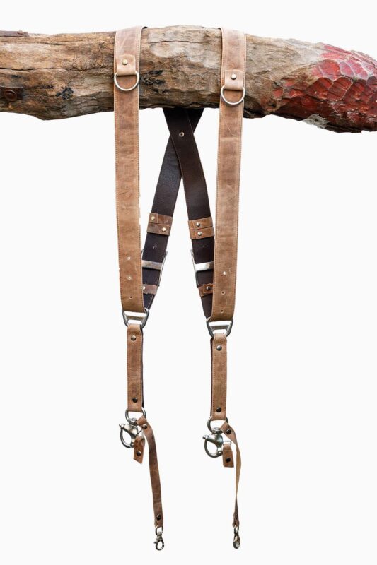 Brown Wild West Vintage leather camera harness displayed on weathered wood log, showing full X-harness design with tan leather shoulder straps, black textured center strap with brown leather accent bands, metal D-ring connectors, and carabiner clips