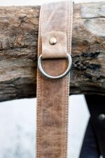 Close-up detail of Brown Wild West Vintage harness shoulder attachment showing metal D-ring, tan vintage leather strap with white stitching, rivet hardware, and natural leather patina