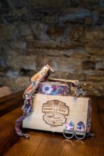 The Flora printed leather camera harness spilling out of the branded bamboo "Genuine Strap - Vintage Design" presentation box on a dark wood table against a rustic stone wall, with two matching floral leather D-ring attachment tabs placed in front of the box.
