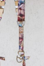 Close-up detail of a single strap end showing the "GENUINESTRAP" branded triangular D-ring connector, floral crackle leather with rivets, the snap-hook camera attachment, and a small floral leather safety tab at the bottom.