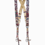 Full-length view of the Flora printed leather dual camera harness hanging from a dark wooden beam, featuring a colorful floral pattern in pink, purple, teal, and gold tones with a cream leather crossback, silver buckles, and snap-hook attachments.