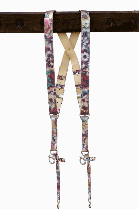 Full-length view of the Flora printed leather dual camera harness hanging from a dark wooden beam, featuring a colorful floral pattern in pink, purple, teal, and gold tones with a cream leather crossback, silver buckles, and snap-hook attachments.