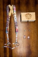 Flat-lay overhead shot of the complete Flora printed leather harness kit: the harness laid out in an X configuration showing the floral print and cream leather backing, the branded bamboo "Genuine Strap - Vintage Design" box, two tripod mounting screws, and two small floral leather D-ring attachment tabs on a dark wood surface.