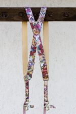 Close-up of the X-shaped crossback junction of the Flora harness hanging from a dark wooden beam, showcasing the vibrant floral print with daisies, roses, and leaves in purple, pink, yellow, and teal on a shimmering textured leather, with gold-tone buckles and cream leather backing straps.