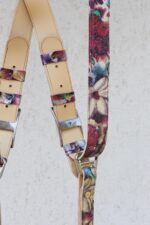 Detail view of the upper portion of the Flora harness showing the cream leather shoulder padding with eagle-embossed branded silver buckles, floral printed leather keeper loops, and the "GENUINESTRAP" engraving on the hardware, alongside the wide floral strap.