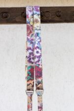 Close-up of both shoulder straps running parallel over a dark wooden beam, highlighting the rich floral print with large yellow blossoms, purple flowers, and teal leaves on the shimmering crackle-textured leather, with branded "GENUINESTRAP" hardware visible below.