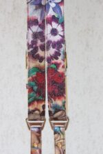 Tight close-up of both harness straps side by side, showing the detailed floral pattern with purple anemones, red roses, and green foliage on crackle-finish leather, silver buckles, and "GENUINESTRAP" branded D-ring connectors at the bottom.