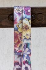 Extreme close-up of both straps draped over a dark wooden beam, showcasing the fine detail of the floral print with large yellow blossoms, pink petals, and purple clematis on the shimmering crackle leather surface, with cream leather backing visible between the straps.
