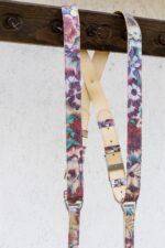 Three-quarter view of the Flora harness hanging from a dark wooden beam, showing the cream leather X-shaped crossback with rivet, branded silver buckles, floral printed keeper loops, and the colorful straps descending toward the D-ring connectors.