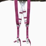 Fuchsia candy leather dual camera harness hanging from a rustic wooden branch against a white background, showing the full front view with two pebbled leather shoulder straps, white leather back panel with leather keepers, silver adjustable buckles, snap shackle clips, and lobster claw clasps at each end.