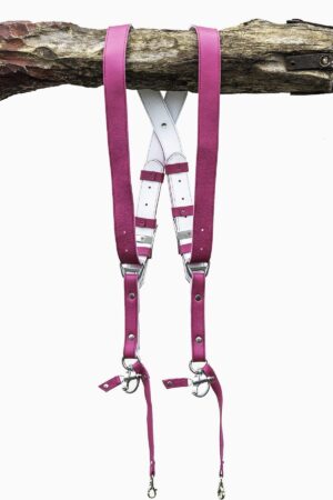 Fuchsia candy leather dual camera harness hanging from a rustic wooden branch against a white background, showing the full front view with two pebbled leather shoulder straps, white leather back panel with leather keepers, silver adjustable buckles, snap shackle clips, and lobster claw clasps at each end.
