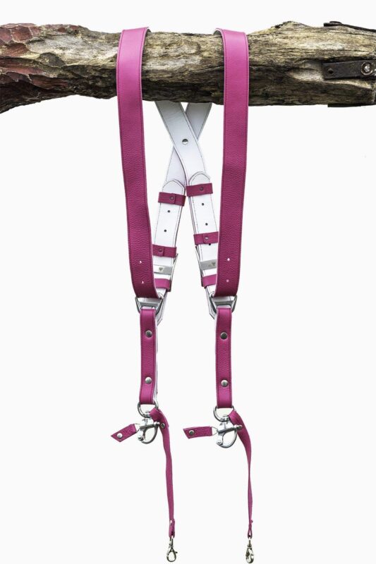 Fuchsia candy leather dual camera harness hanging from a rustic wooden branch against a white background, showing the full front view with two pebbled leather shoulder straps, white leather back panel with leather keepers, silver adjustable buckles, snap shackle clips, and lobster claw clasps at each end.