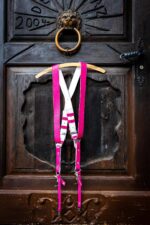 Fuchsia candy leather dual camera harness displayed on a wooden hanger hung from a bronze lion door knocker on an antique dark wooden door, showing the full X-cross back configuration with white leather backing, silver buckles, and snap hook attachments.
