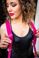 Close-up portrait of a female photographer wearing the fuchsia candy leather camera harness, showing the wide pebbled leather shoulder straps over a black top, with silver hardware visible at the lower edges.