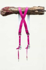 Fuchsia candy leather dual camera harness hanging from a rustic wooden branch against a light background, displayed from the back showing the X-cross strap configuration, silver adjustable buckles, snap shackle clips, and lobster claw clasps at each end.