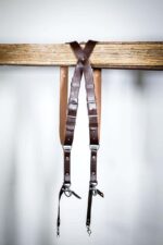 Dark brown leather dual camera harness straps hanging on a wooden rail, full length view.
