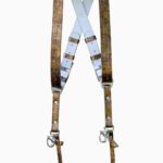 Full view of the gold embossed leather dual camera harness hanging from a rustic iron bar against a white background, showing the complete front view with two wide crinkled metallic gold shoulder straps, white leather back panel with X-cross design, branded "GENUINESTRAP" connectors, adjustable buckles, snap shackle clips, and lobster claw clasps at each end.