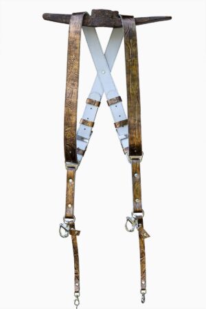 Full view of the gold embossed leather dual camera harness hanging from a rustic iron bar against a white background, showing the complete front view with two wide crinkled metallic gold shoulder straps, white leather back panel with X-cross design, branded "GENUINESTRAP" connectors, adjustable buckles, snap shackle clips, and lobster claw clasps at each end.