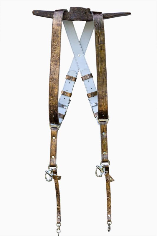 Full view of the gold embossed leather dual camera harness hanging from a rustic iron bar against a white background, showing the complete front view with two wide crinkled metallic gold shoulder straps, white leather back panel with X-cross design, branded "GENUINESTRAP" connectors, adjustable buckles, snap shackle clips, and lobster claw clasps at each end.