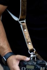 Close-up of a photographer's hand holding a camera at hip level, showing the gold embossed leather harness strap running from shoulder to camera with the branded "GENUINESTRAP" metal connector, silver rivets, D-ring, and snap shackle clip against a dark background.