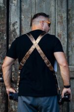 Rear view of a photographer wearing the gold embossed leather dual camera harness over a black t-shirt, showing the X-shaped cross-back design with silver adjustable buckles, two cameras hanging at each hip, standing in front of a weathered green wooden door.