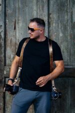 Front view of a photographer wearing sunglasses and the gold embossed leather dual camera harness over a black t-shirt, holding two cameras at his sides with the textured metallic gold straps draped over both shoulders, photographed against a rustic green wooden door.