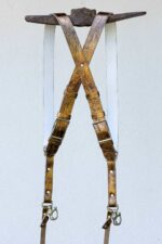 Full view of the gold embossed leather dual camera harness hanging from a rustic iron bar against a light wall, displayed from the back showing the X-cross strap configuration, crinkled metallic gold leather, silver adjustable buckles, branded metal connectors, snap shackle clips, and lobster claw clasps at each end.