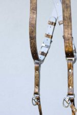 Detail shot of the mid-section of the gold embossed leather camera harness showing both straps with the white leather back panel, branded "GENUINESTRAP" metal connectors, leather keepers, silver rivets, D-rings, and snap shackle clips at the bottom.