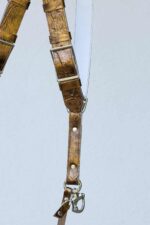 Close-up detail of a single strap junction on the gold embossed leather camera harness, showing the silver adjustable buckle, crinkled metallic gold leather texture with stitching, rivets, D-ring connector, snap shackle clip, and leather safety tether against a light background.