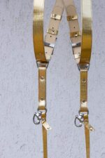 Front-facing symmetrical view of both harness straps hanging side by side, showing the branded silver slide buckles, cream leather padding sections, wave-textured gold leather, rivets, and matching snap hooks at the bottom.