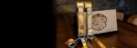 Gold Tingala leather camera harness with metallic wave-textured straps, silver hardware, and snap hooks, displayed leaning against a branded wooden "Genuine Strap - Vintage Design" presentation box on a dark wood surface.