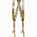 Full-length view of the Gold Tingala leather dual camera harness hanging from a dark wooden beam, showing the X-shaped crossback design, adjustable silver buckles, cream leather backing, and two snap-hook attachment points at the bottom.