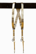 Full-length view of the Gold Tingala leather dual camera harness hanging from a dark wooden beam, showing the X-shaped crossback design, adjustable silver buckles, cream leather backing, and two snap-hook attachment points at the bottom.