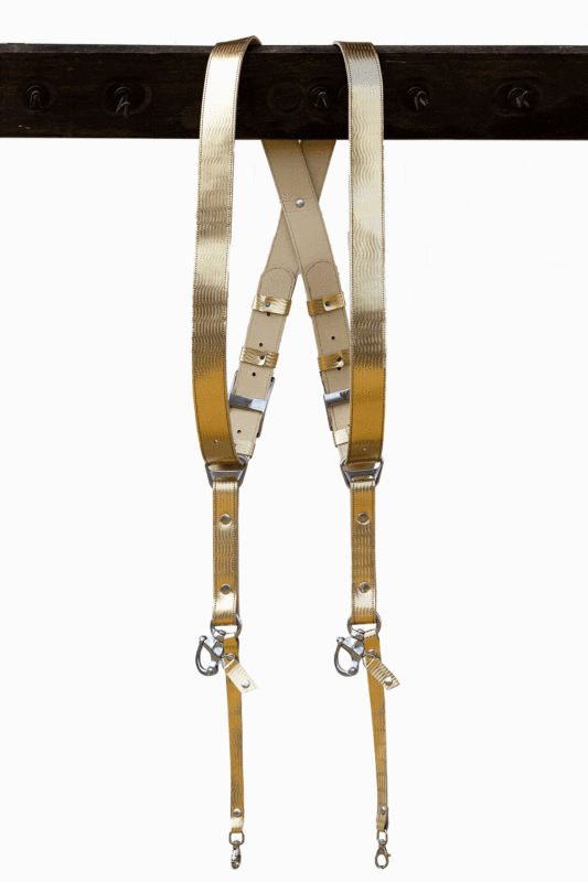 Full-length view of the Gold Tingala leather dual camera harness hanging from a dark wooden beam, showing the X-shaped crossback design, adjustable silver buckles, cream leather backing, and two snap-hook attachment points at the bottom.