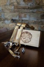 Gold Tingala camera harness draped over the branded bamboo presentation box on a dark wood table, with loose snap hooks and a small metal mounting screw visible beside it, set against a rustic stone wall.