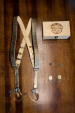 Flat-lay overhead shot of the complete Gold Tingala harness kit: the full harness laid out in an X configuration, the branded bamboo box, two tripod mounting screws, and two small leather D-ring attachment tabs, all arranged on a dark wood surface.