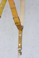 Close-up detail of the Gold Tingala harness showing the adjustable silver buckle, wave-textured gold leather strap with stitched edges, cream leather lining, metal rivets, and the snap-hook camera attachment at the bottom.
