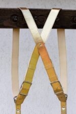 Close-up of the X-shaped crossback junction of the Gold Tingala harness hanging from a dark wooden beam, highlighting the rivet at the crossing point, dual silver buckles, and cream leather backing straps.