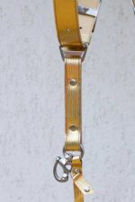 Detail view of a single strap end showing the branded silver slide buckle with eagle emblem, herringbone-textured gold leather, metal rivet, D-ring, and marine-grade snap hook with a leather safety tab.