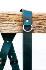 Close-up of Green Amulet harness showing two-tone leather (dark and teal), metal D-ring attachment point, and white stitch detailing on connector flap