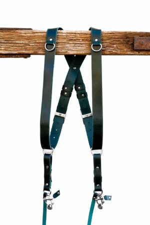 Green Amulet leather camera harness showing dual-tone teal and dark green leather, full X-harness design with adjustable segments and metal connectors over wooden beam