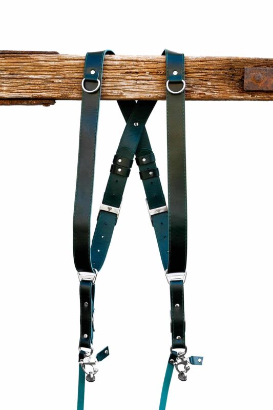Green Amulet leather camera harness showing dual-tone teal and dark green leather, full X-harness design with adjustable segments and metal connectors over wooden beam